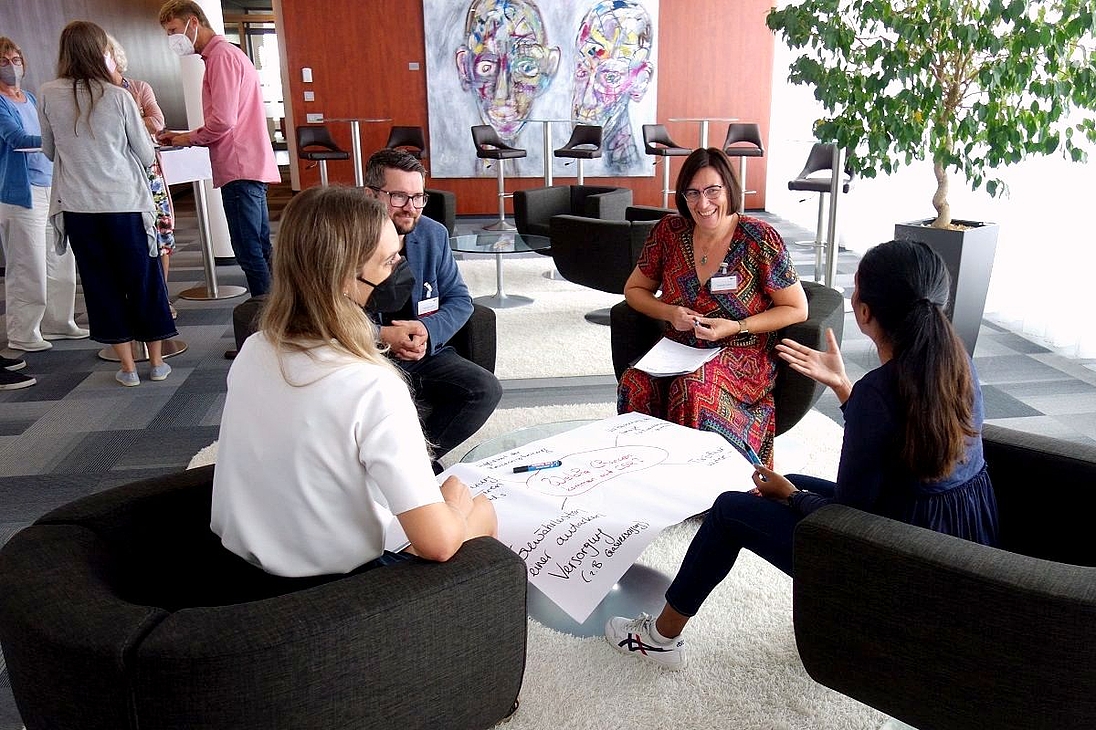 iga.Expertendialog 2022: Zwei Kleingruppen befinden sich im intensiven Austausch. Eine Gruppe sitzt um einen Tisch und hat ein beschriebenes Flipchart-Blatt in ihrer Mitte liegen. Die andere Gruppe steht im Hintergrund um einen Stehtisch.