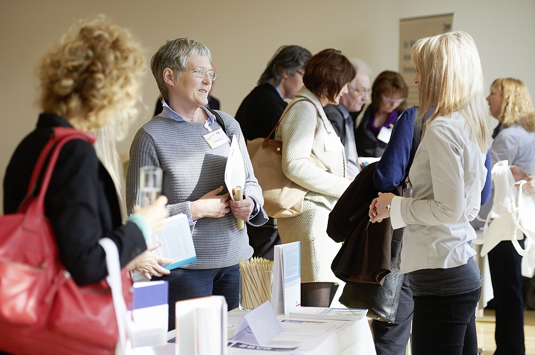 iga.Kolloquium 2015: Auf einem Tisch liegt Informationsmaterial aus. Im Hintergrund diskutieren Teilnehmende.