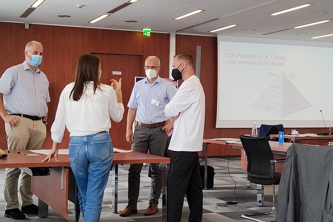 iga.Expertendialog 2022: Eine Gruppe aus vier Personen dikutiert eine Fragestellung. Im Hintergrund ist eine Folie zu sehen, auf der anhand einer Pyramide das amerikanische Modell der CSR nach A. B. Carroll dargestellt ist.