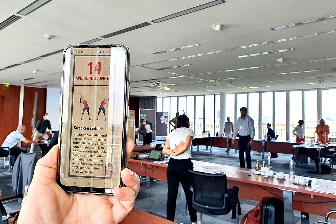 iga.Expertendialog 2022: Ein Teilnehmender leitet eine bewegte Pause an. Er nutzt dafür ein digitales Tool, das im Vordergrund des Bildes auf einem Smartphone eingeblendet ist. Alle führen jeweils die Übung aus, die auf ihrem Smartphone angezeigt wird.