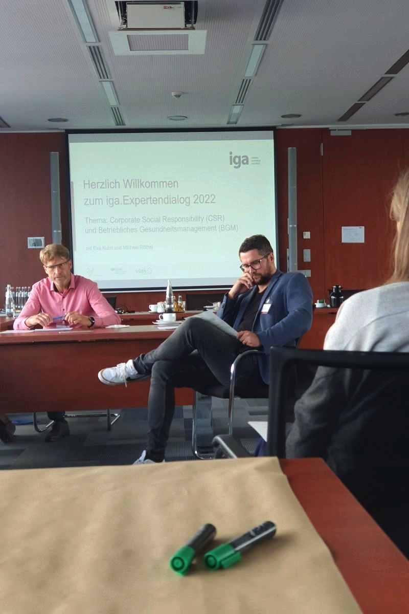 iga.Expertendialog 2022: Teilnehmende denken über eine Fragestellung nach. Im Hintergrund ist eine Folie mit dem Titel der Veranstaltung zu sehen: Corporate Social Responsibility (CSR) und Betriebliches Gesundheitsmanagement (BGM).
