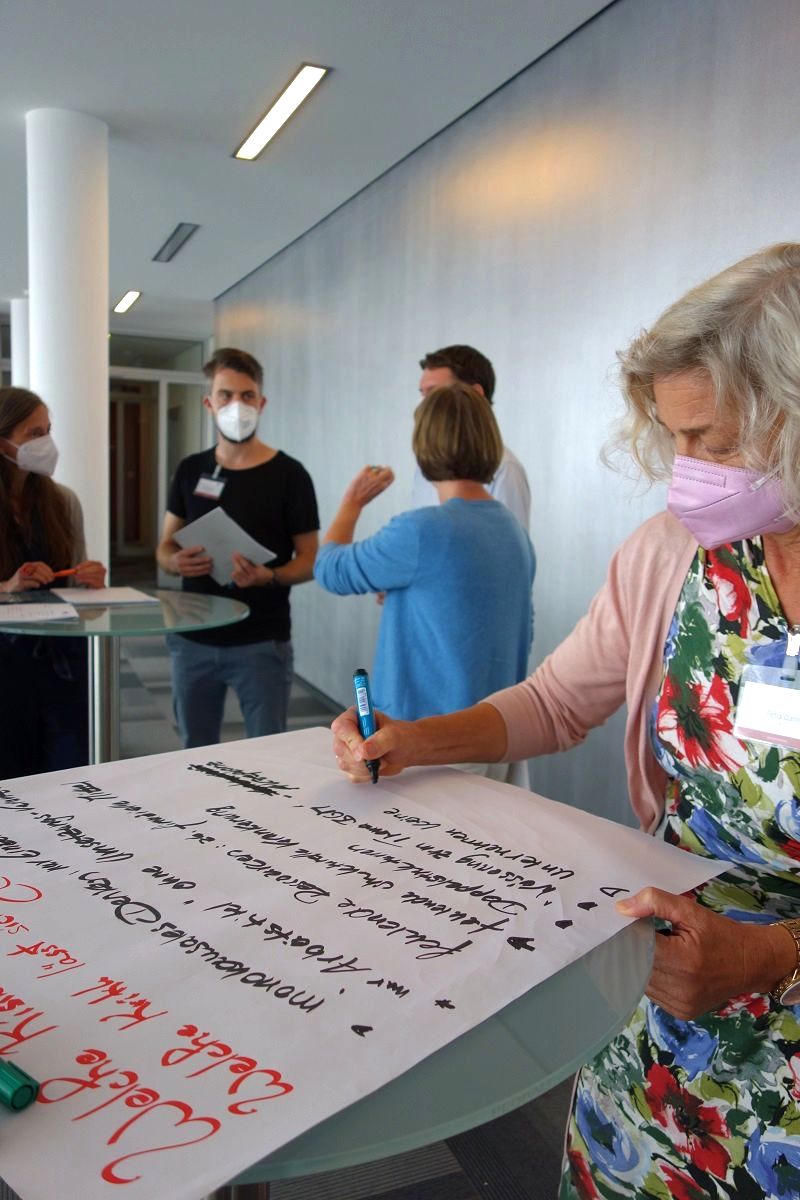 iga.Expertendialog 2022: Eine Teilnehmerin notiert auf einem Flipchart-Blatt Aspekte zu einer Fragestellung, die auf dem Blatt rot notiert ist. Im Hintergrund tauschen sich ihre Kolleginnen und Kollegen weiter aus.