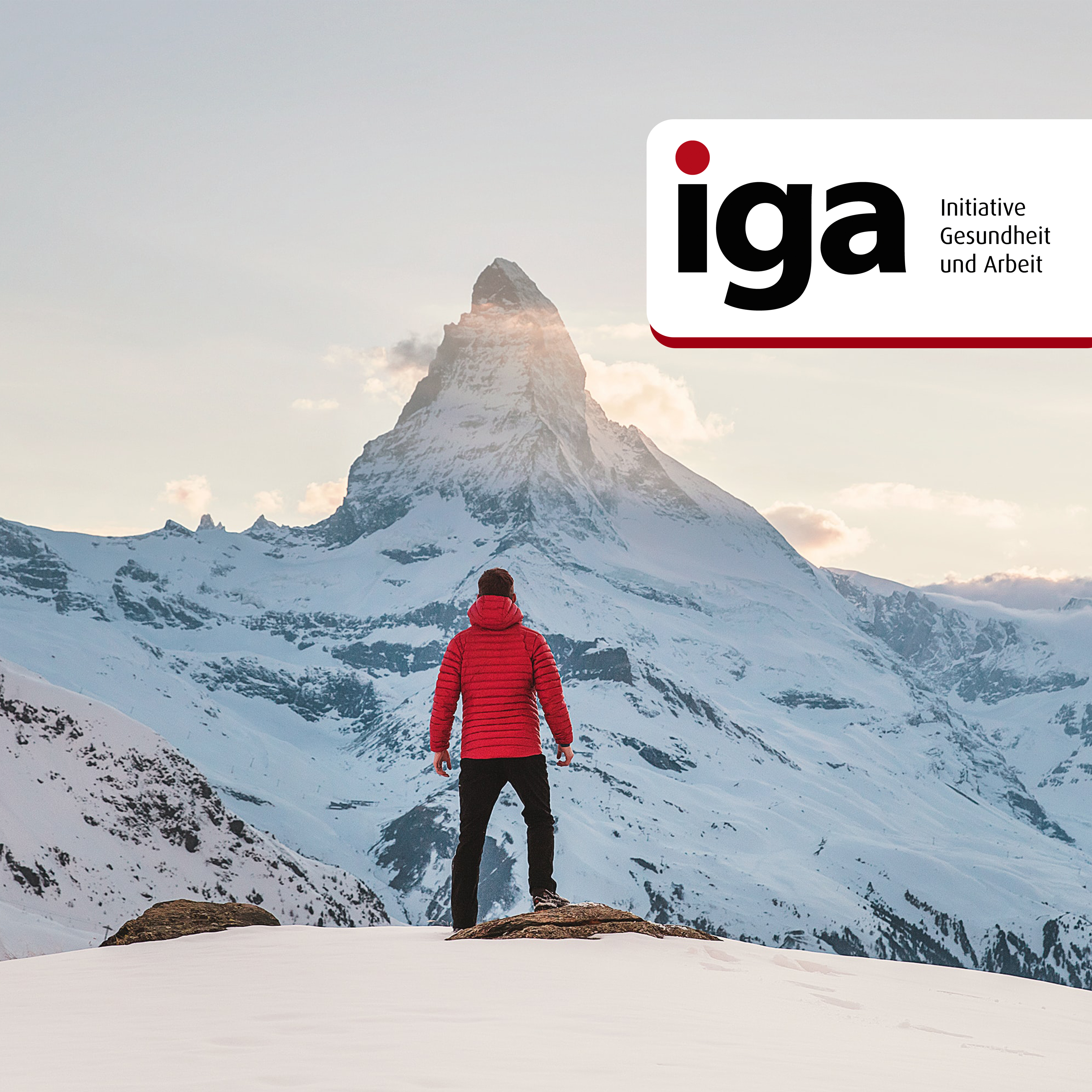 Ein Mann in roter Winterjacke steht vor einem hohen Berg. Er blickt zielstrebig zur Bergspitze hinauf..