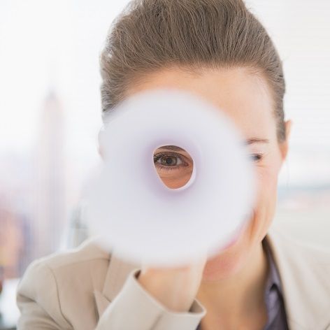 Eine Frau blickt durch ein zum Fernrohr gerolltes Blatt Papier.