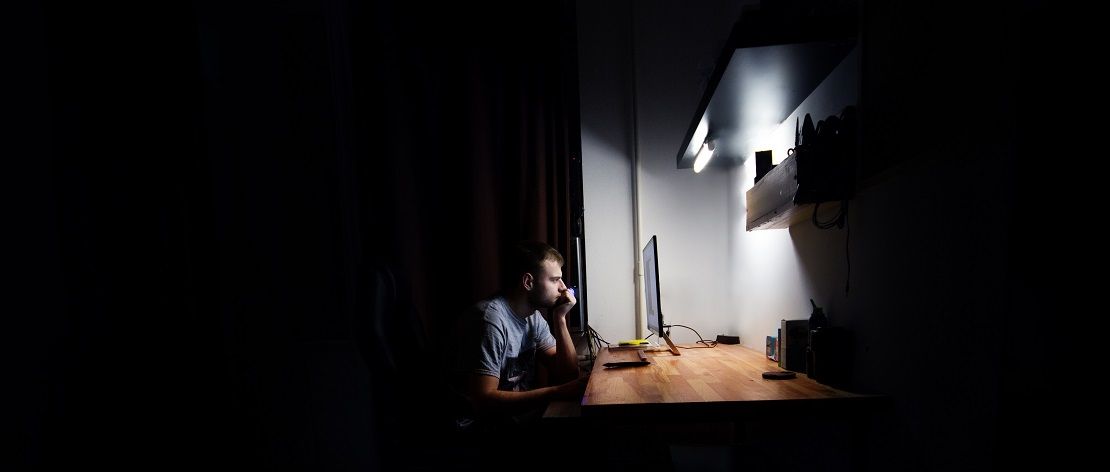 Ein junger Mann sitzt am heimischen Schreibtisch allein und im Dunkeln vor dem Computer.