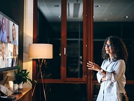 Eine Chefin hält mit ihrem Team eine Videokonferenz ab. Bei ihr ist es bereits abends und dunkel. Die 6 Teammitglieder sind auf einem großen Wandbildschirm zu sehen. 