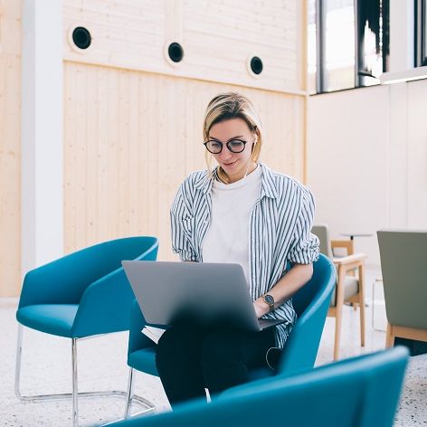 Eine junge Frau sitzt auf einem Sessel und arbeitet an ihrem Laptop. Sie befindet sich in einem großen, hellen Arbeitsraum. 